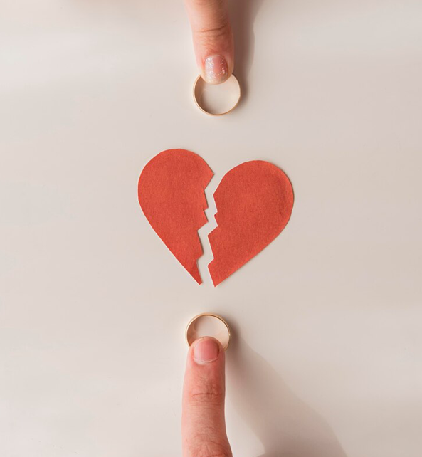 A female finger holding a ring, a red broken heart, and male finger holding a ring.