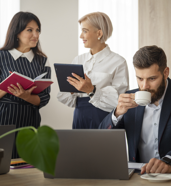 Two female lawyer Fort Pierce professionals are standing and one attorney Fort Pierce professional man sitting in front of his laptop, sipping his coffee.